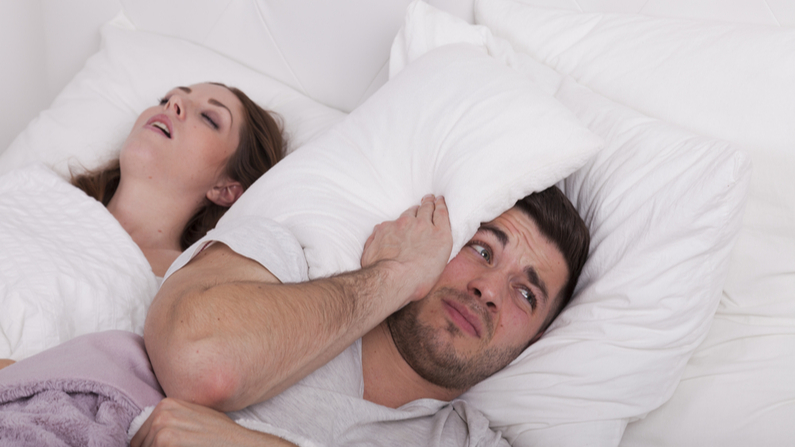 Woman snoring in bed, while the man covers his ears.